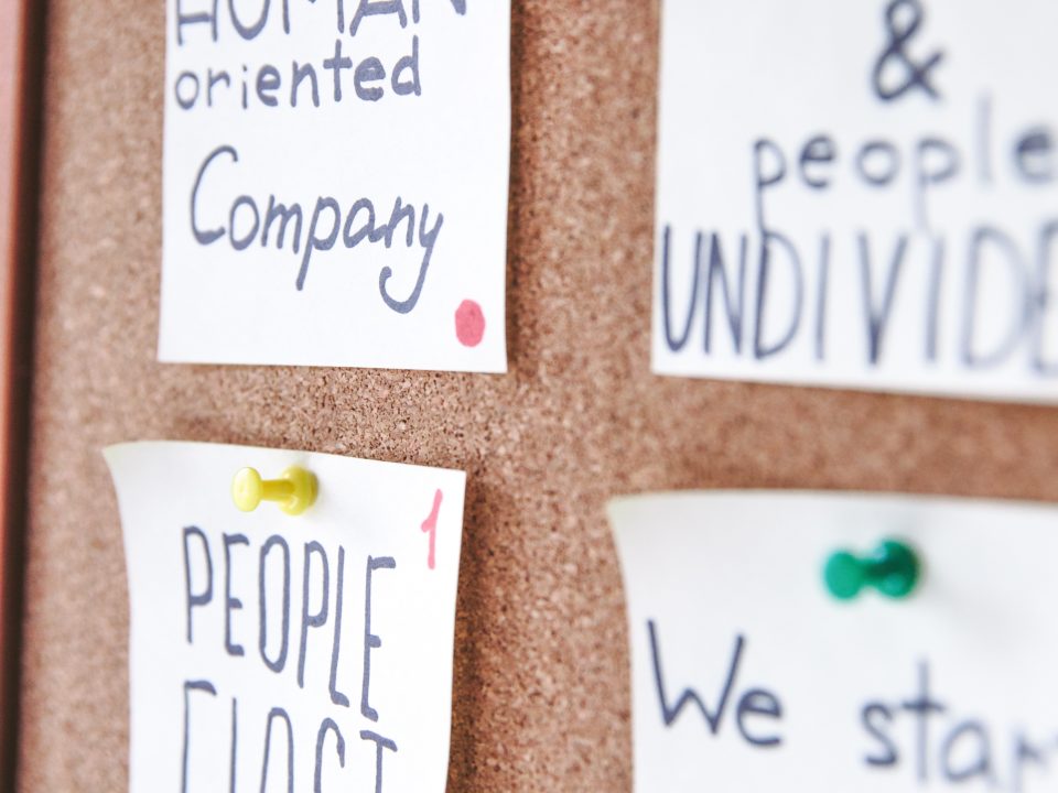 Photo of a cork board with hand written cards affixed with push pins that say, "Human-Oriented Company" and "People First"